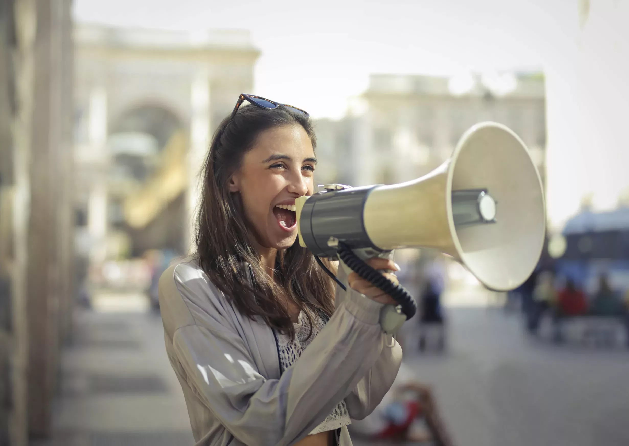A woman with long dark hair wearing a beige jacket and sunglasses on her head is holding a bullhorn and smiling widely, conveying a sense of positivity and enthusiasm.