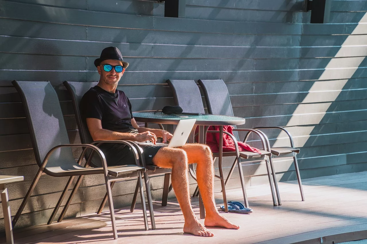 Man sitting outdoors at the pool on a lounge chair, working on a laptop while enjoying the sun